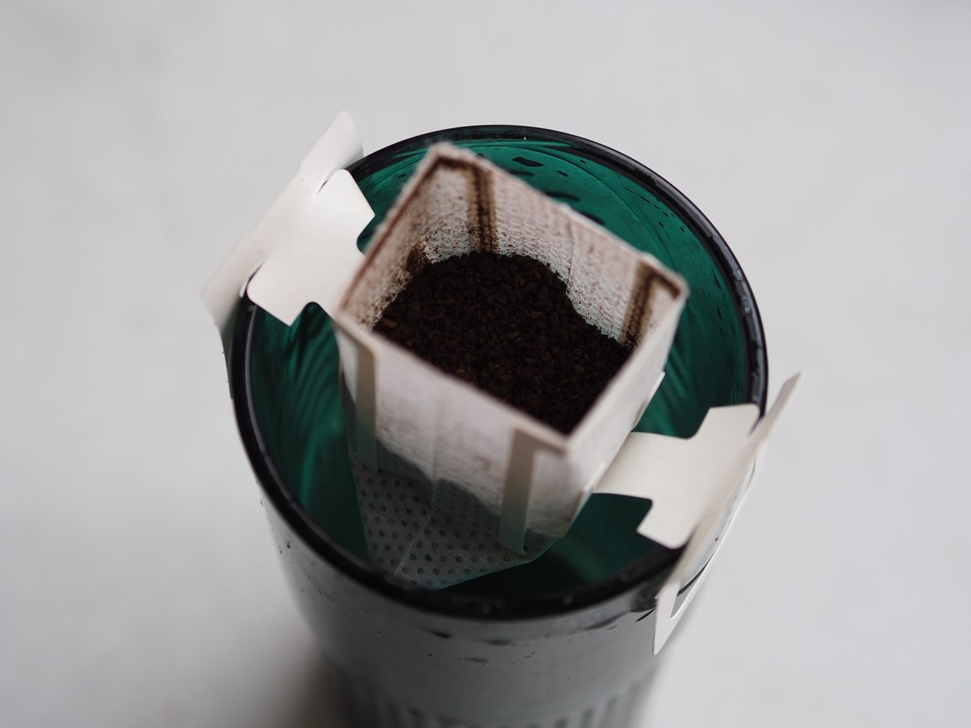 A single-serve drip coffee bag with ground coffee, hooked over the rim of a mug