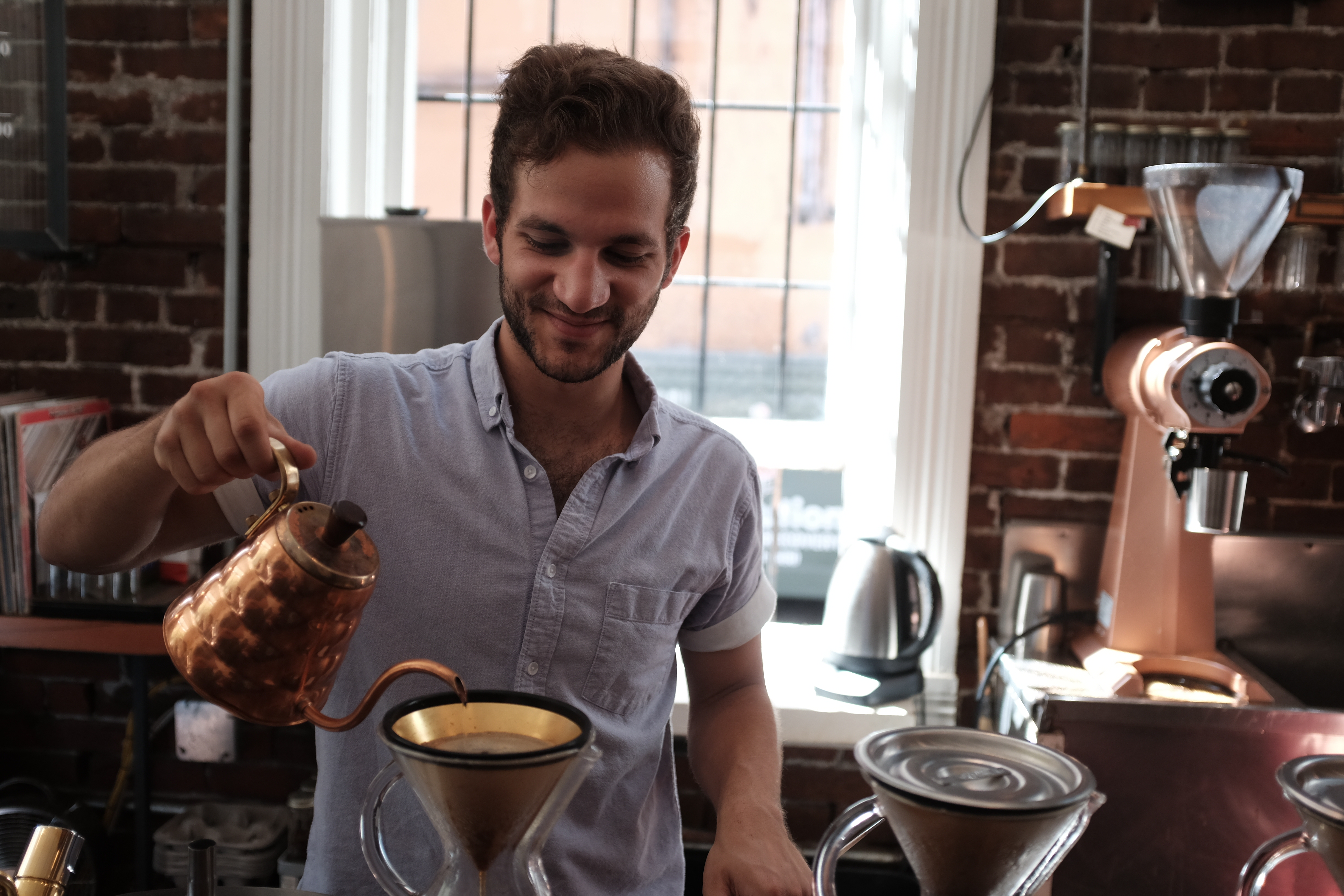 A person pouring water from a gooseneck kettle over a cone dripper in a café