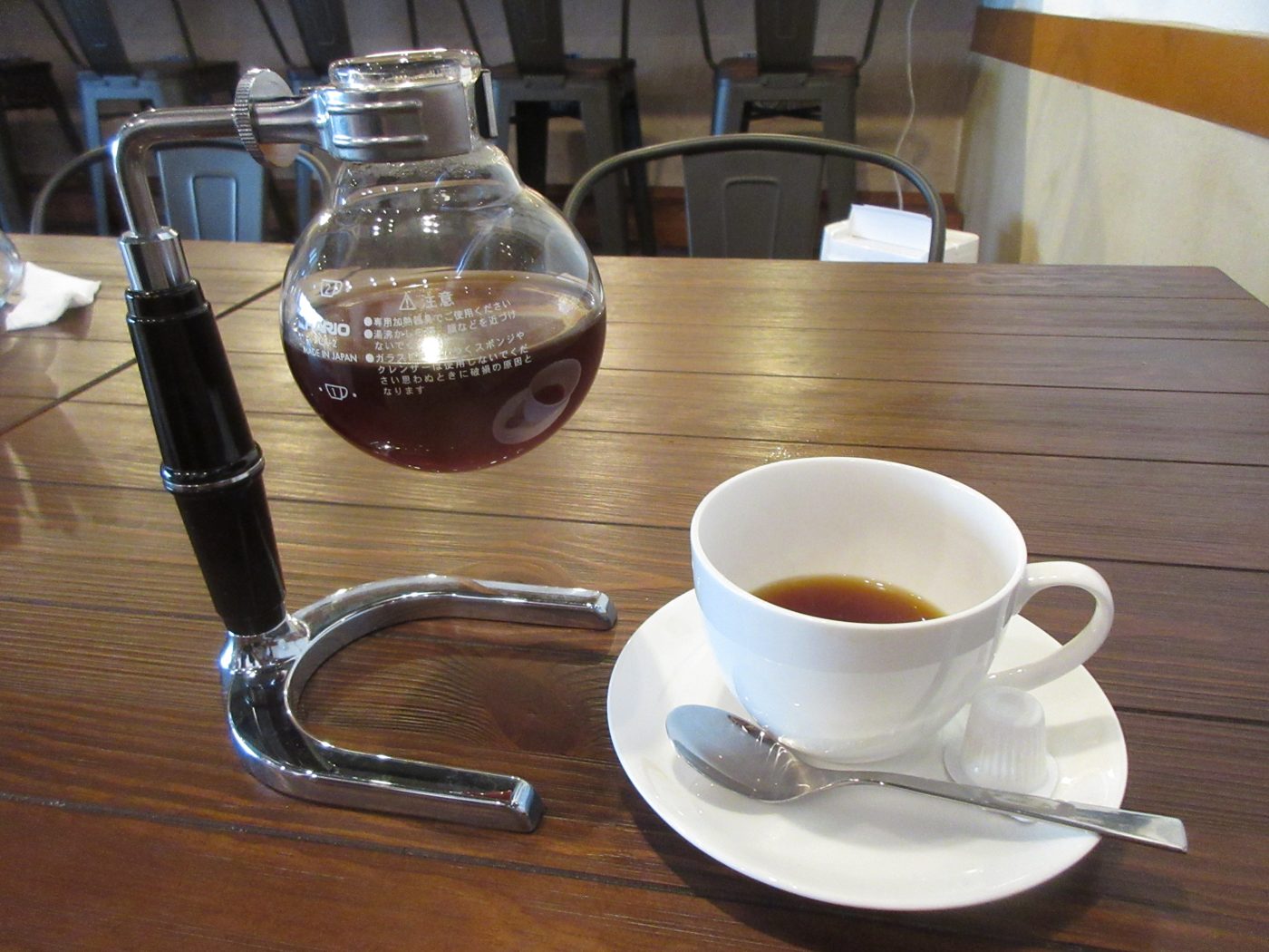 A glass vacuum coffee maker with its upper chamber, filter and stand, next to a served cup